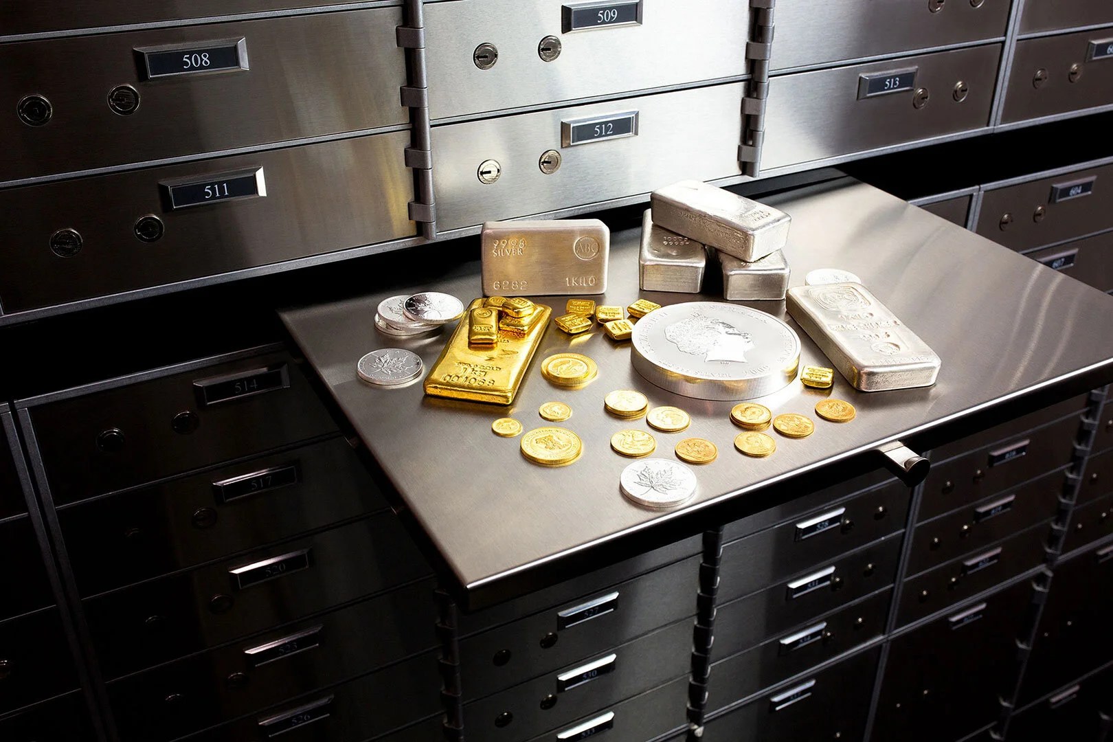 Gold and silver bullion and coins laid out on the tray of a safety deposit box drawer, with numbered boxes behind.